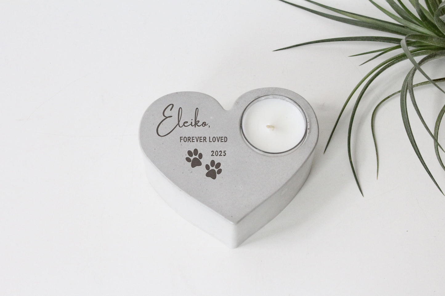 A heart-shaped concrete candle holder with a candle inside, placed on a white surface next to a plant.