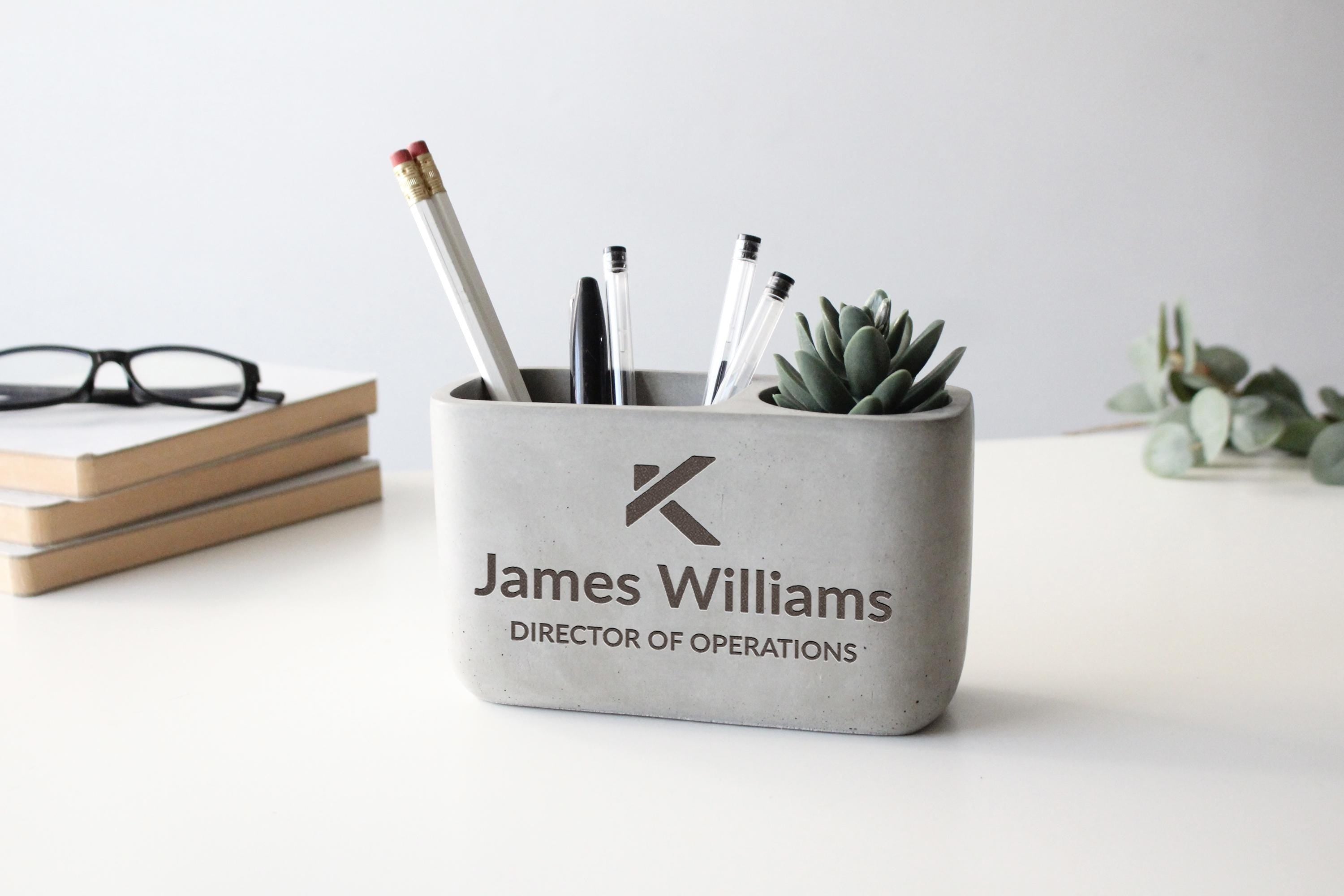 A gray container holding pens, pencils, and a small succulent plant, with the text "James Williams DIRECTOR OF OPERATIONS" printed on it.