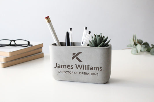 A gray container holding pens, pencils, and a small succulent plant, with the text "James Williams DIRECTOR OF OPERATIONS" printed on it.