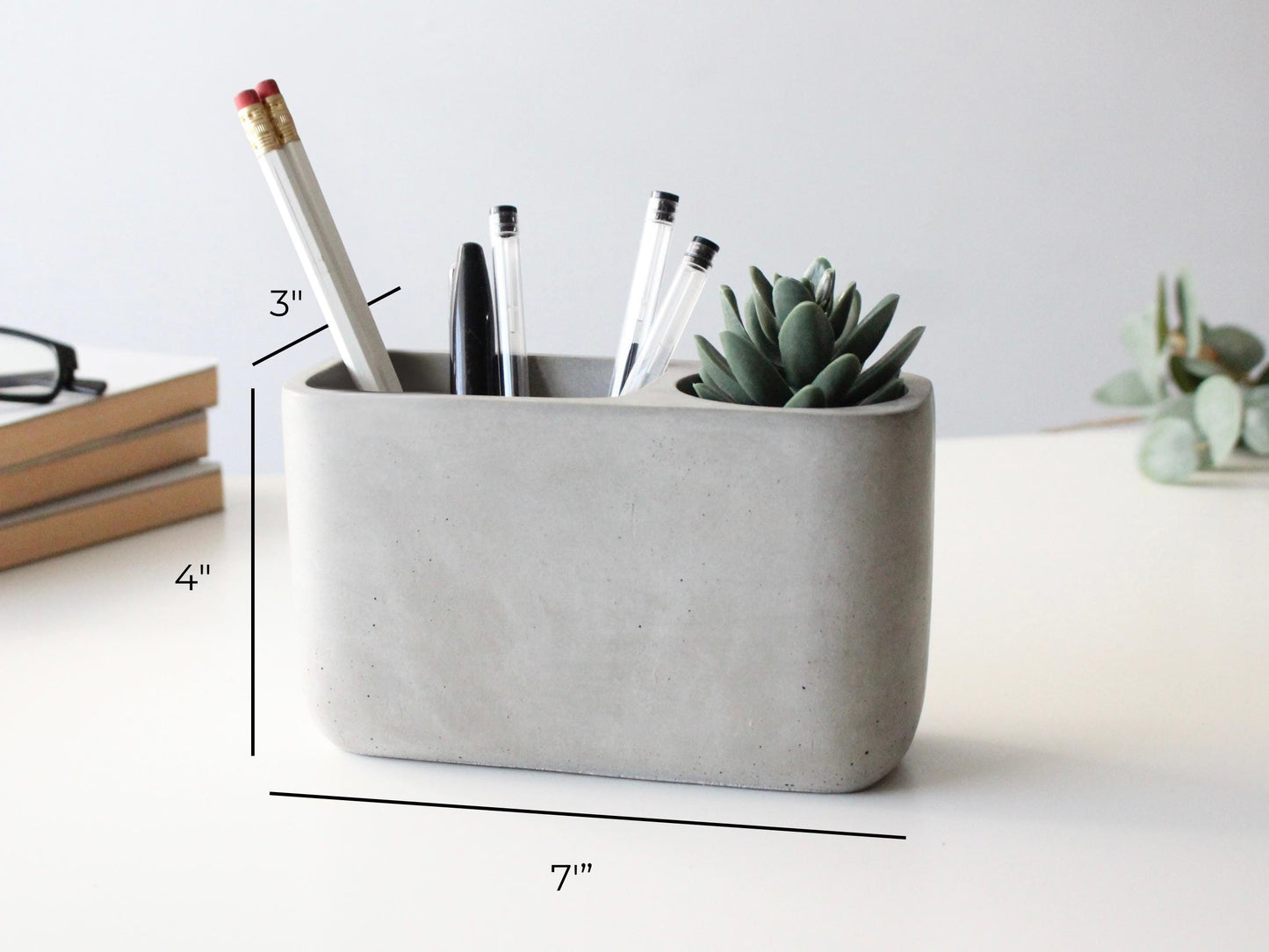 A gray concrete pencil holder with pens and a succulent plant in it, placed on a white surface.