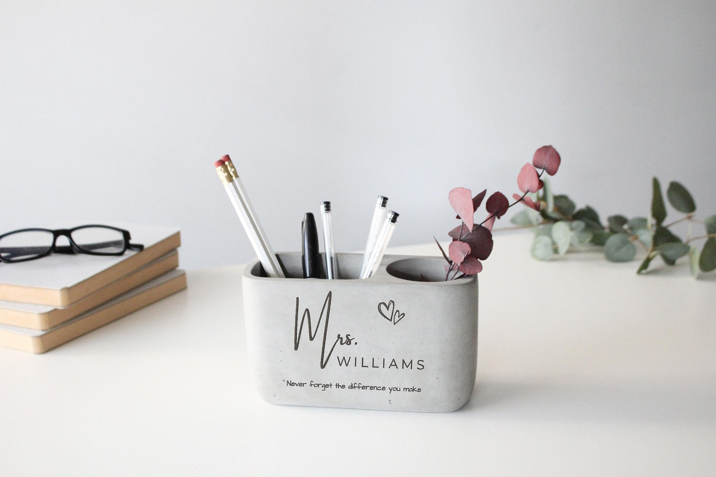 A gray pencil holder on a white desk, containing pens and pencils, with a pair of glasses and a stack of books nearby.