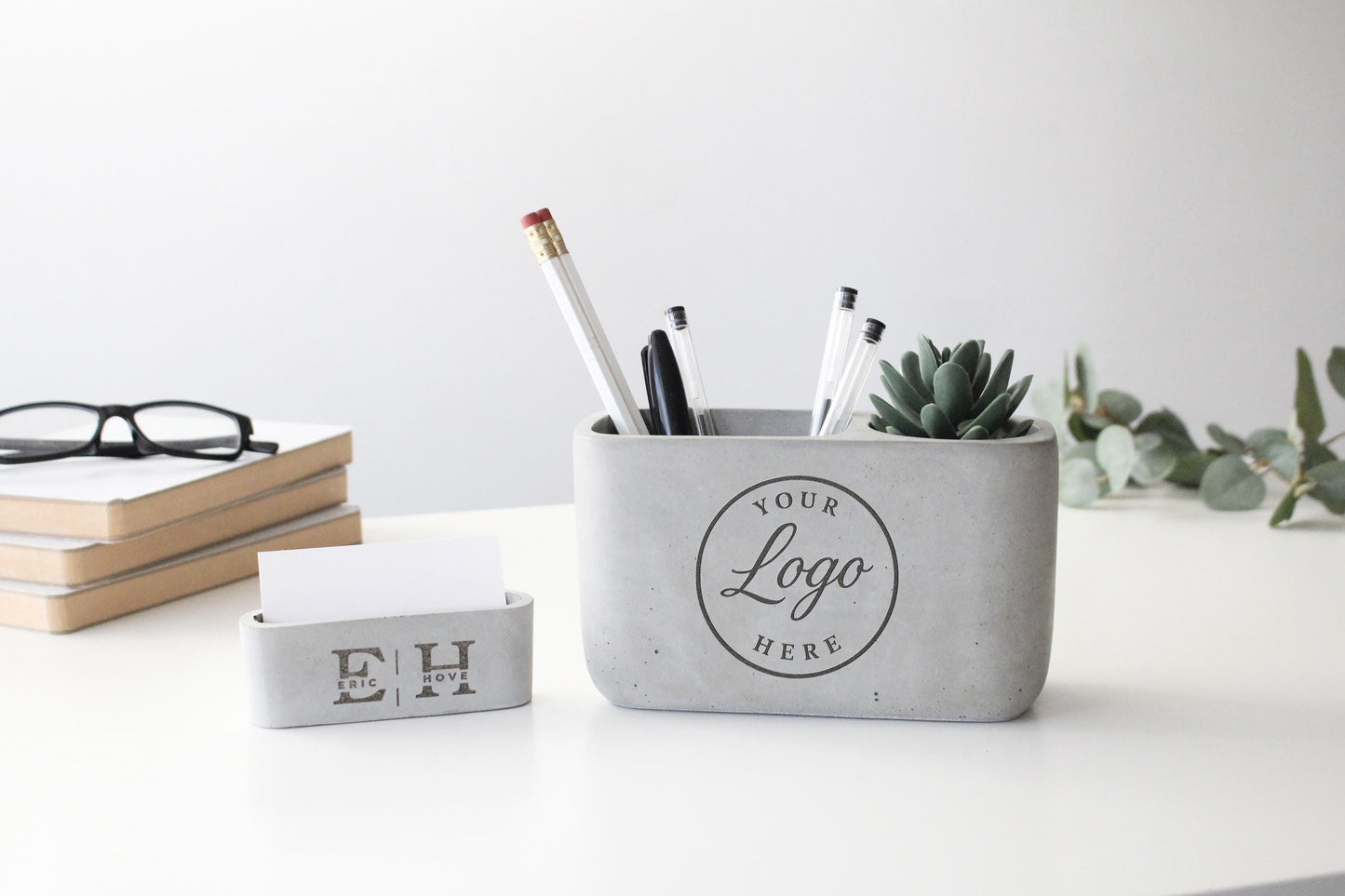 a gray container with a logo, a pen holder, and a small white card holder on a white desk.