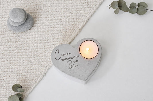 A heart-shaped concrete candle holder with a candle inside, placed on a white surface. The candle holder has a personalized message engraved on it.