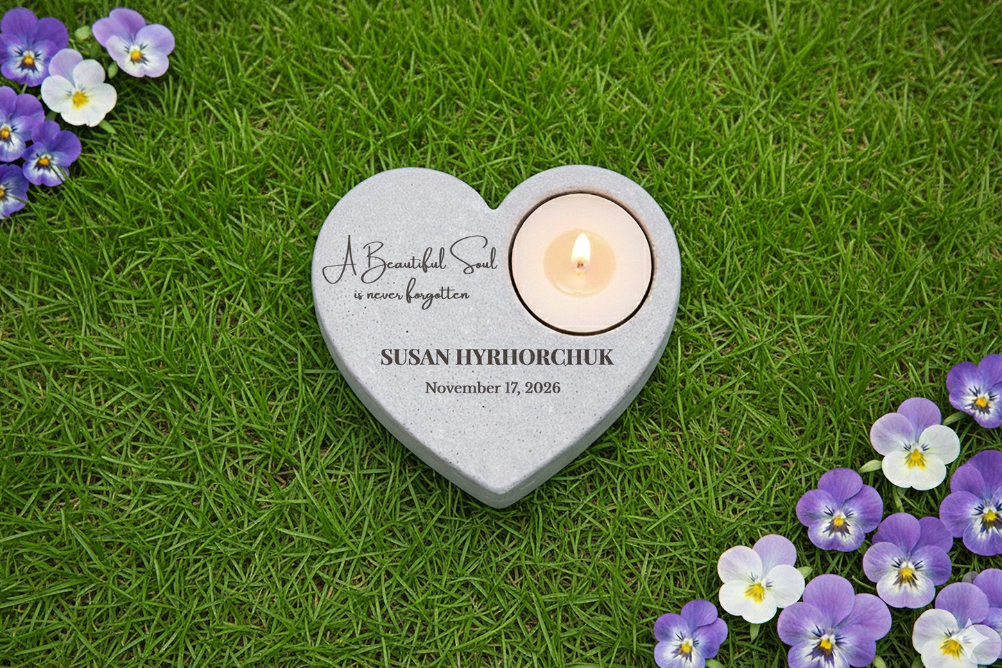 A heart-shaped stone with a lit candle in it, surrounded by purple and white flowers, on a grassy surface.