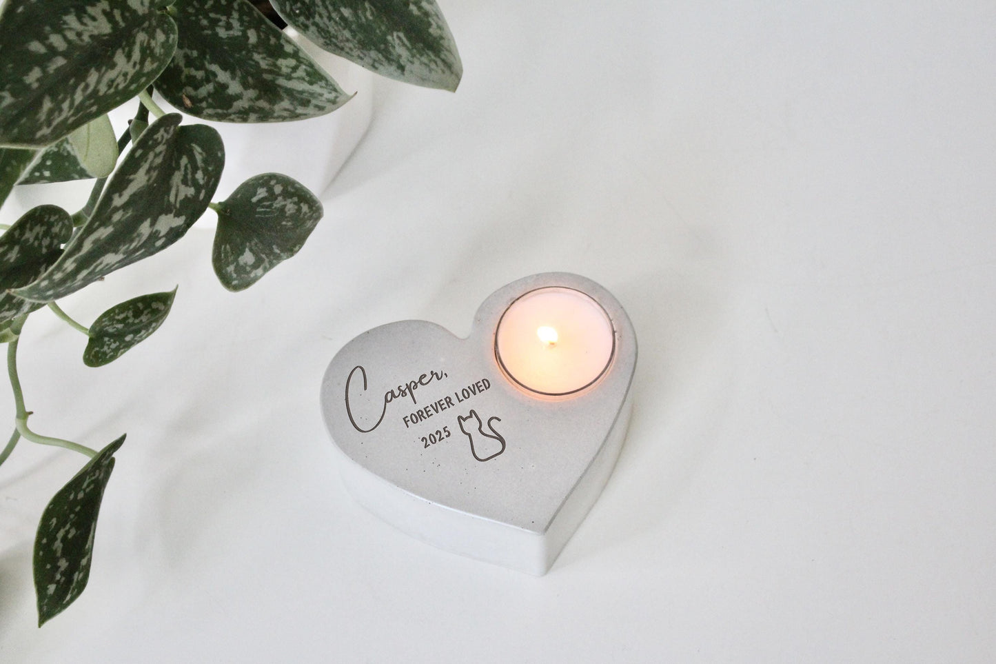 A heart-shaped candle holder with a lit candle inside, placed on a white surface next to a potted plant.